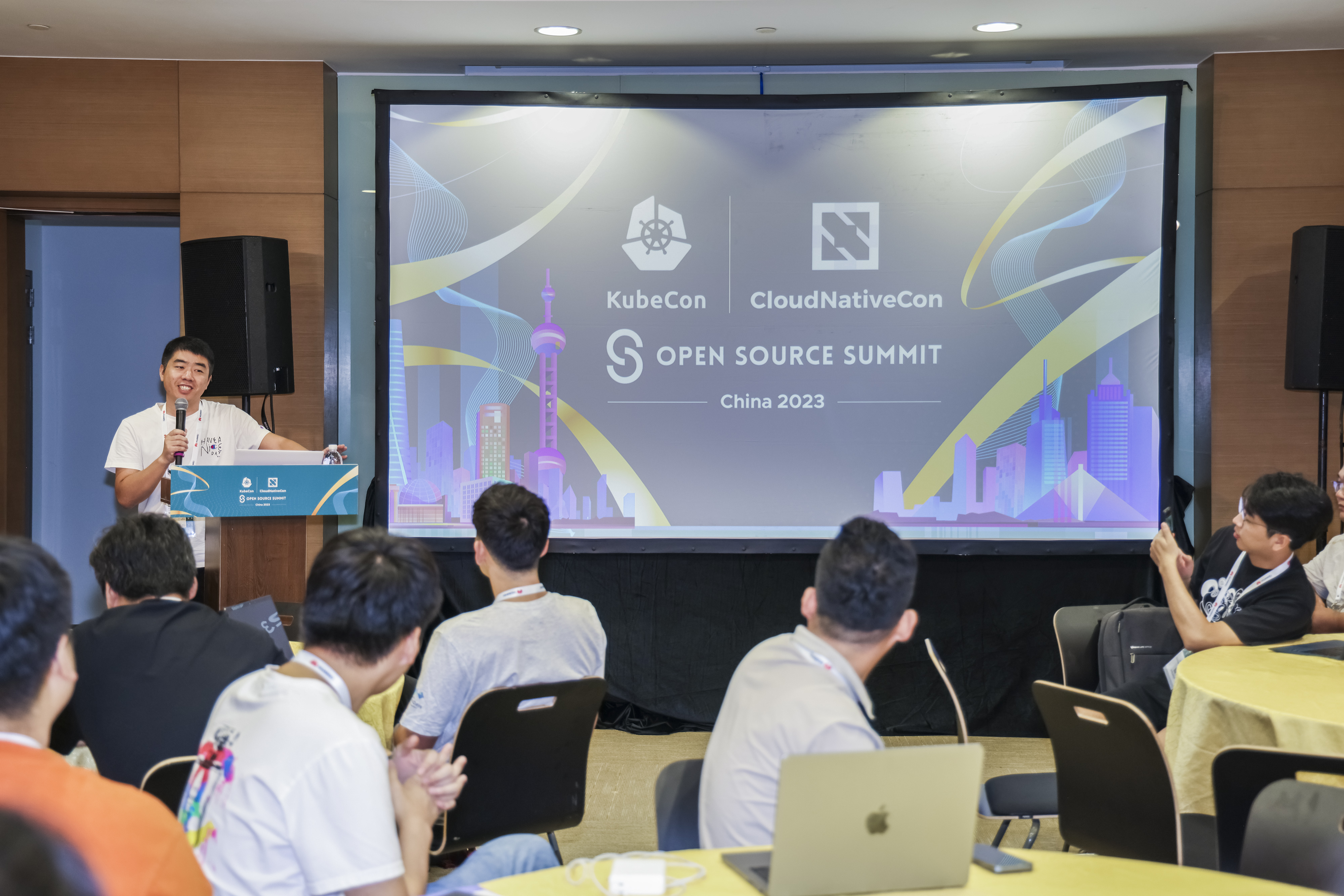 A group of contributors sat at tables, listening to a presenter speaking at a podium